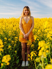 Naklejka na ściany i meble Portrait of cheerful woman on rapeseed field with sunset light. Yellow flowers and happy woman