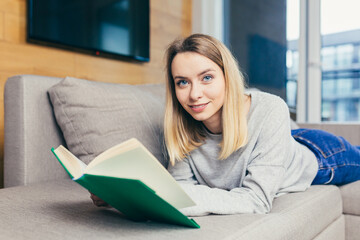 Young beautiful blonde woman reads an interesting book while lying on the couch, resting after work