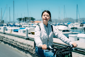 carefree asian woman is looking into the distance while enjoying riding a bike along marina with sailboats in Sausalito city, California on a sunny vacation day