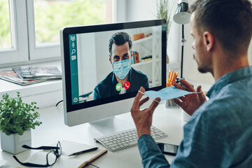Confident young man in protective face mask talking to collegue by video call
