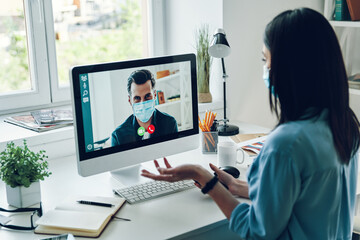 Serious young woman in protective face mask talking to collegue by video call