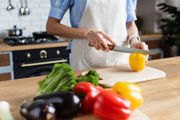 partial view of young adult woman cutting yellow pepper on cutting board in kitchen
