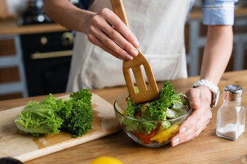 partial view of young adult woman mixing vegetables salad with spatula in kitchen