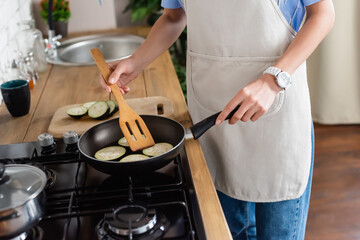 cropped view of young adult woman frying slices of eggplant in pan and using spatula in kitchen