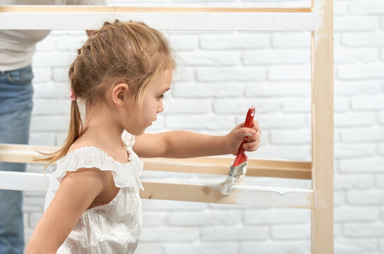 Pretty little child with blond hair painting wooden shelves with brush and white color. Cute kid helping mother to restoring furniture at home.