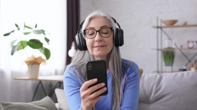 Happy Beautiful Elderly Gray-haired Woman In Black Headphones Enjoy Listening Favorite Music On Smartphone Using Music App Sitting On The Couch At Home.Relax,stress Management.Good Start To The Day.