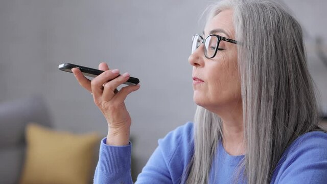 Smiling elderly woman recording voice message on smartphone. Attractive senior lady using virtual assistant app sitting at home. Voice recognition, audio messages, using translator on mobile phone.
