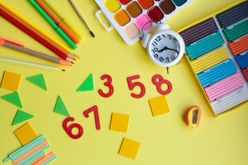School background with school supplies on yellow, pen, pencils, markers, watercolors, plasticine, sharpener, numbers, geometric shapes, counting sticks, alarm clock, plasticine, flat lay