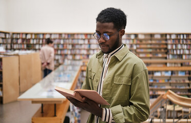 African young man in eyeglasses reading a book while standing in the library