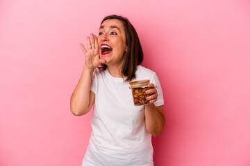 Middle age caucasian woman holding an almond jar isolated on pink background shouting and holding palm near opened mouth.