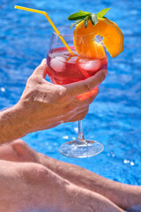 Man at the edge of a swimming pool holding a very appetizing cocktail with one hand