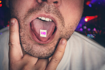 guy swallows puts a brand of narcotic acid on his tongue against background of a concert in a club. Junkies have fun at night on raver party at a nightclub. Close-up on junkie young man is taking LSD. - obrazy, fototapety, plakaty