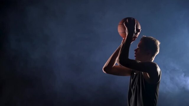 A young basketball player in a black sports uniform makes a jump shot, side view. The guy trains before the competition in a dark, smoky studio under the spotlights. Slow motion. Close up.