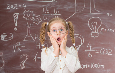 Funny shocked  little girl in glasses near chalkboard full of signs and formulas. Education concept.