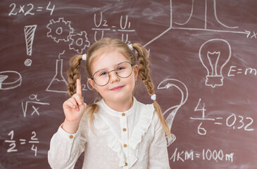 Funny smiling  little girl in glasses near chalkboard full of signs and formulas. Education concept.