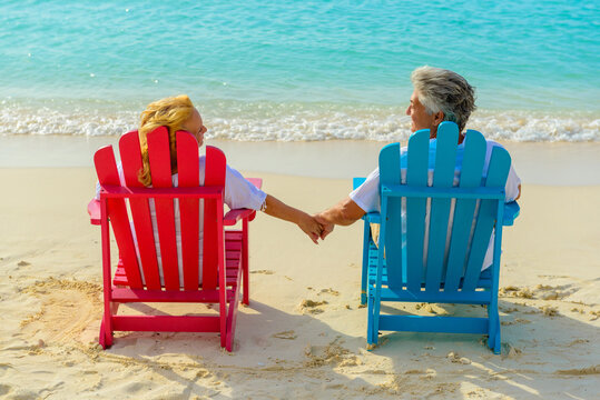 Older Couple At The Beach Holding Hands And Sitting On Beach Chairs