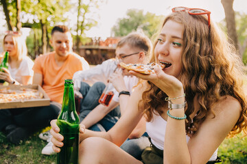 Young curly girl eating pizza and drinking beer together with her friends outdoors
