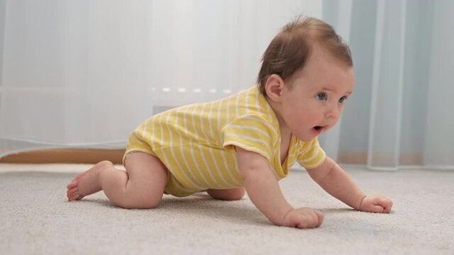 Early development of child. Cute little child in yellow clothes is lying on floor in nursery, crawling on his hands on carpet of house. First steps of child on way to crawling. Child learns to crawl.