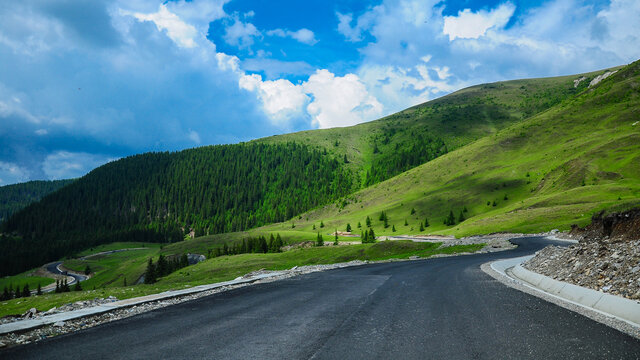 Transalpina road winding along an alpine grassland near rocky mountain ridges and forested mountain peaks. Clouds cast their shadows on the grassy meadows. Carpathia, Romania.