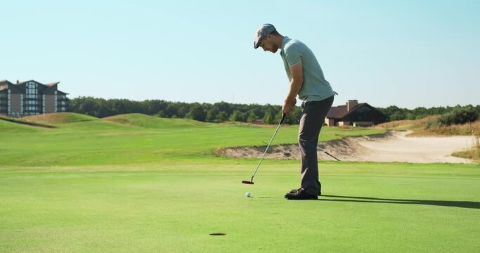 Caucasian male golfer wearing cap and golf clothes, swinging and hitting ball into hole, playing at green lawn