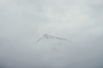 Minimal mountain landscape with high pointy rock in clouds. Minimalist mountain scenery with sharp snowy mountain peak over clouds. Snow-white pointed pinnacle above white clouds. Big top in dense fog