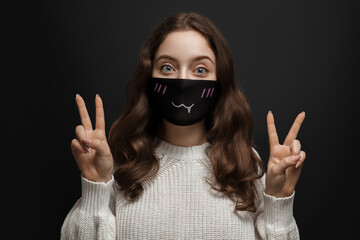 A young girl in a mask against a dark background shows a gesture of peace