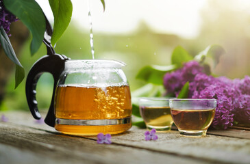 A glass teapot with hot herbal tea flowing and glass bowls on a wooden table against a background...