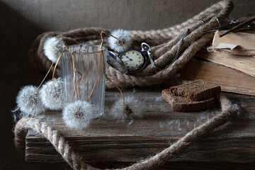 Vintage still life with ripe dried dandelions in a faceted glass and an old pocket watch on an old wooden table.
