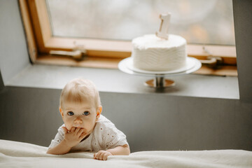 Fototapeta premium Front view of little female child standing near a window. There is a birthday cake on the window cell.
