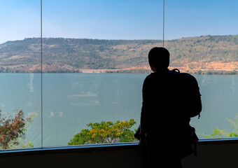 silhouette back of backpack tourist man standing and looking at damn and mountain view at transparent window from indoor room, Lamtakong dam view observation in Nakhon Ratchasima, Thailand