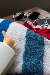 Blue and white striped towel, cream bottle, sunglasses, black smartphone, earphones and red sort pants for summer on a brown wooden table 