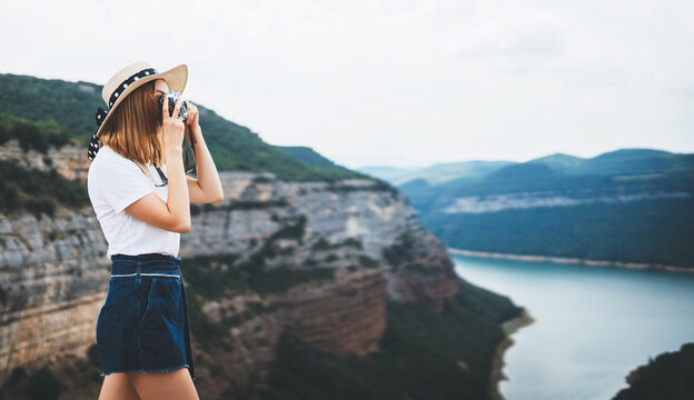 young blonde girl in summer hat takes photo on retro camera on background panorama river horizin mountain landscape, hipster tourist enjoys hobby of photographer  leisure in summer nature empty space