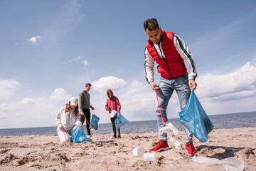 young man holding trash bag and collecting rubbish on sand near volunteers