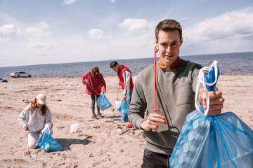 happy man holding trash bag near group of blurred volunteers picking up rubbish on sand © LIGHTFIELD STUDIOS