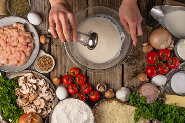 In the photo we see the process of preparing the dough. The cook in the blender mixes the ingredients for the dough. Bright vegetables and herbs lie on a wooden table. View from above.
