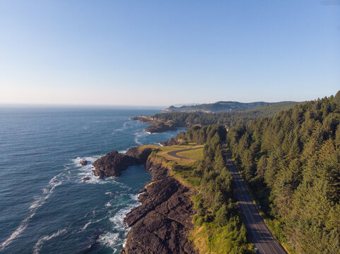 In the photo, you see a highway near the ocean. On the left side is the ocean, rocky coast. On both sides of the road there are hills overgrown with green grass. View from above.
