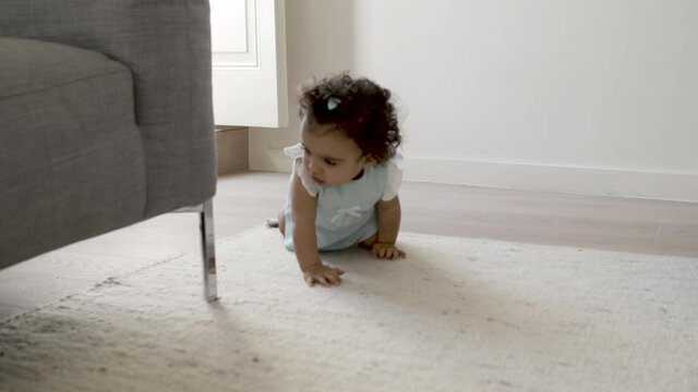 Front view of cute focused infant crawling on carpet. Little girl sitting on floor, doing efforts to stand up with help of sofa. Crawling, childhood concept