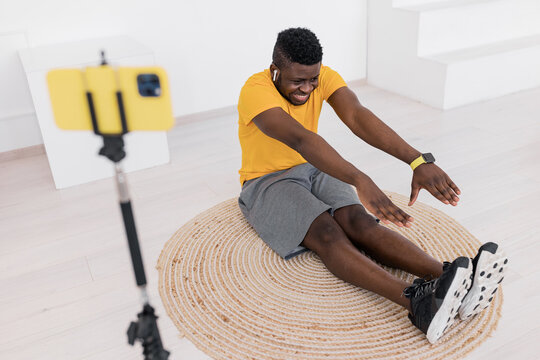 Smiling African American man coach in yellow t-shirt practicing yoga at online lesson,doing Seated forward bend exercise, using smart phone on tripod to hold online tutorial at home during quarantine