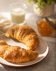 In the photo we see a still life of two ruddy croissants on a white plate, golden orange jam and a glass of milk. Pastel shades. White background. Bright lighting. There are no people in the photo.