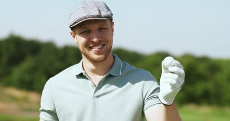 Young caucasian man in cap wearing professional gloves holding golf ball and smiling widely to camera at golf course