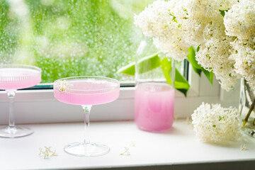 Obraz na płótnie Canvas Fresh pink non alcoholic cocktail in glass with lilac flowers against window background.