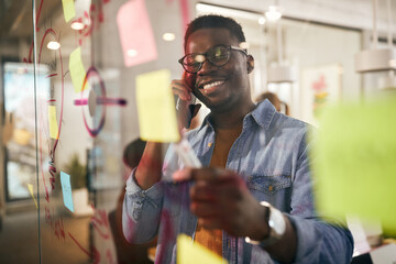 Happy black businessman talking on the phone while analyzing mind map on glass wall in the office.