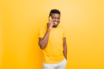 Portrait of an attractive smiling trendy young african american man looking at camera, empty copy space, isolated over yellow background, talking on mobile phone with friends