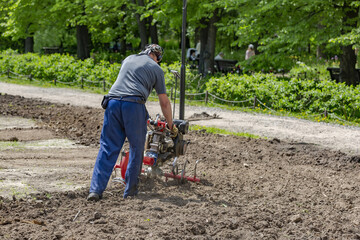 Tall Caucasian man in uniform with a tiller plows the soil in a green garden
