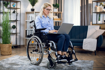 Fototapeta na wymiar Pleasant young woman with disabilities working on modern laptop while staying at home. Charming blonde in eyewear and casual clothes typing on keyboard while sitting in wheelchair.