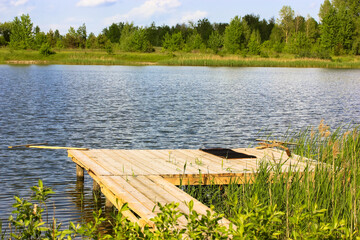 Jetty in calm lake, river. A natural summer landscape. An old wooden boardwalk in a swampy area near water body. Location for fishing and recreation in a nature reserve. Tall grass, trees and reeds.