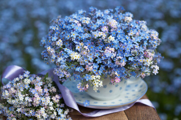 A bouquet spring of forget-me-nots in a cup and on a table on a blurred background of blue flowers outdoor. Blur, selective focus, postcard. - Powered by Adobe