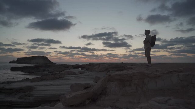 Sea sunset view. Man with backpack on rocks with beautiful view of Yeronisos Island near coast of Agios Georgios Pegeias. Guy enjoying ocean horizon, panoramic sunset in cyprus on mediterranean sea