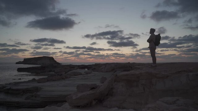 Man with backpack is hiking along a rocky coast at sunset near the Mediterranean sea in Cyprus. Male tourist admires the view Yeronisos, or Holy Island, lies off the coast of Agios Georgios Pegeias