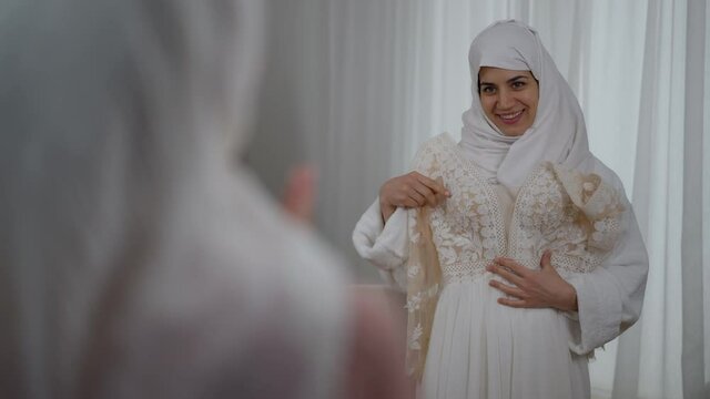 Portrait of happy young Middle Eastern bride with toothy smile trying on wedding dress with excited blurred bridesmaid talking gesturing at front. Joyful women discussing stylish clothes indoors